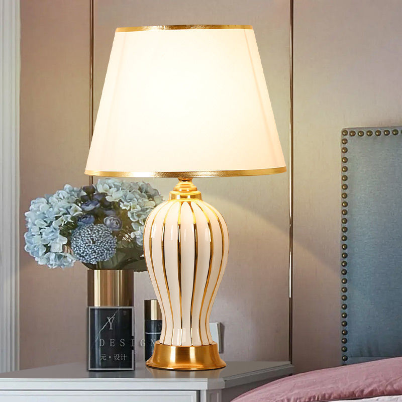 White ceramic table lamp with gold trim on a bedside table with hydrangea flowers, lit