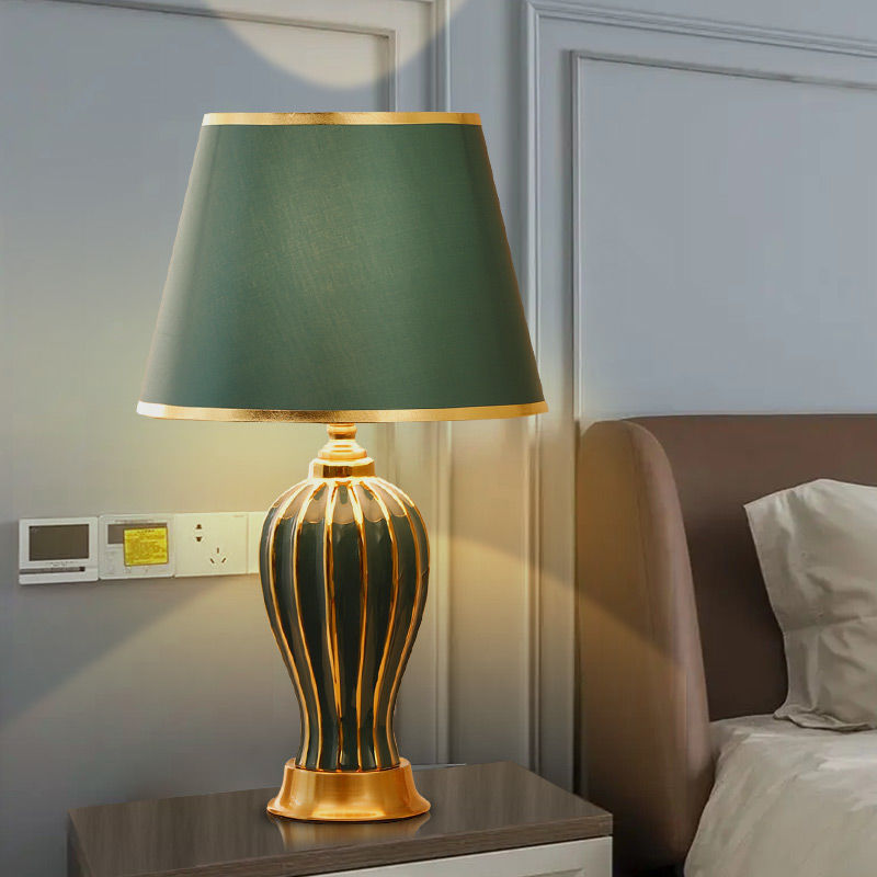 Green ceramic table lamp with fabric shade on a bedside table beside a bed