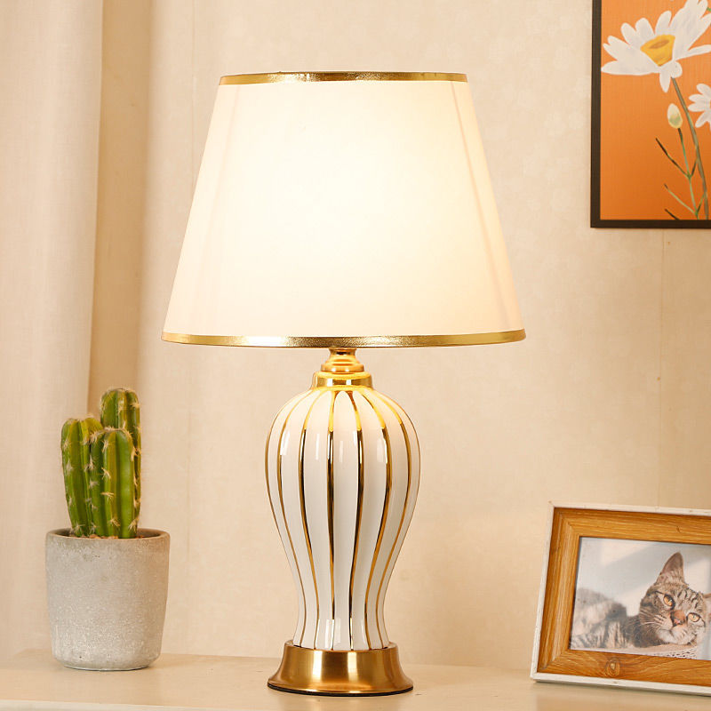 White ceramic table lamp with gold-striped base, lit, with cactus and framed photo