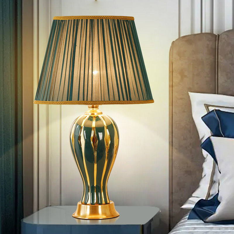 Green ceramic table lamp with pleated shade on a bedside table, lit