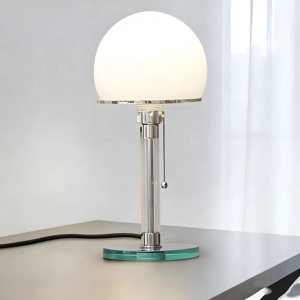 A modern table lamp with a glowing white dome shade, a polished silver metal column, and a round translucent aqua glass base, shown on a tabletop against a light gray wall and white curtain.