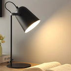 A modern black metal table lamp with a conical shade and adjustable stand, illuminated to cast a warm glow on a beige wall above an open book on a wooden desk.