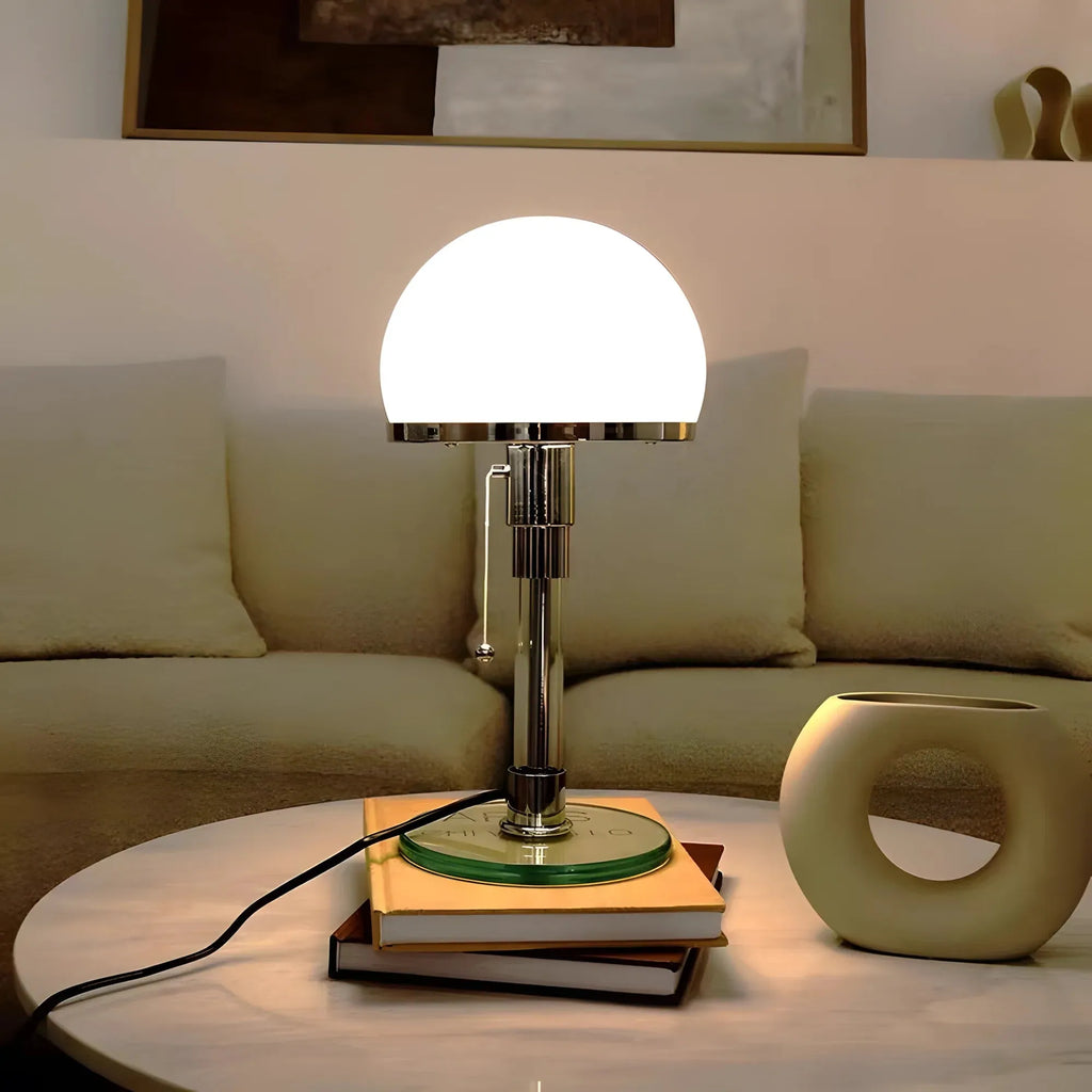 A modern table lamp with a glowing white dome shade and a polished silver metal stem sits on a stack of books on a round white table in a living room setting. A beige sofa and a sculptural ceramic vase are visible in the background.