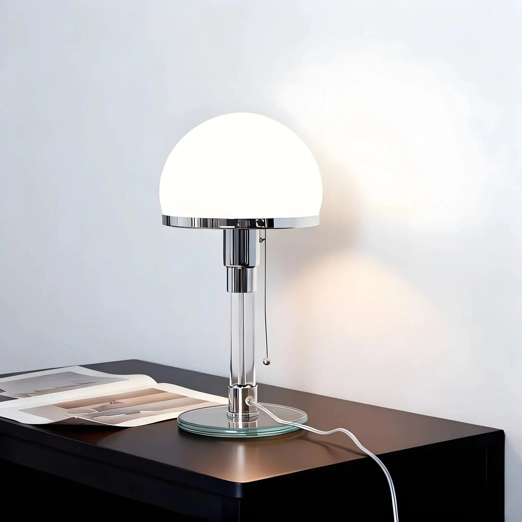 A modern table lamp with a glowing white dome shade and a polished silver metal stem, sitting on a dark wooden side table against a light gray wall.