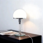 A modern table lamp with a glowing white dome shade and a polished silver metal stem, sitting on a dark wooden side table against a light gray wall.