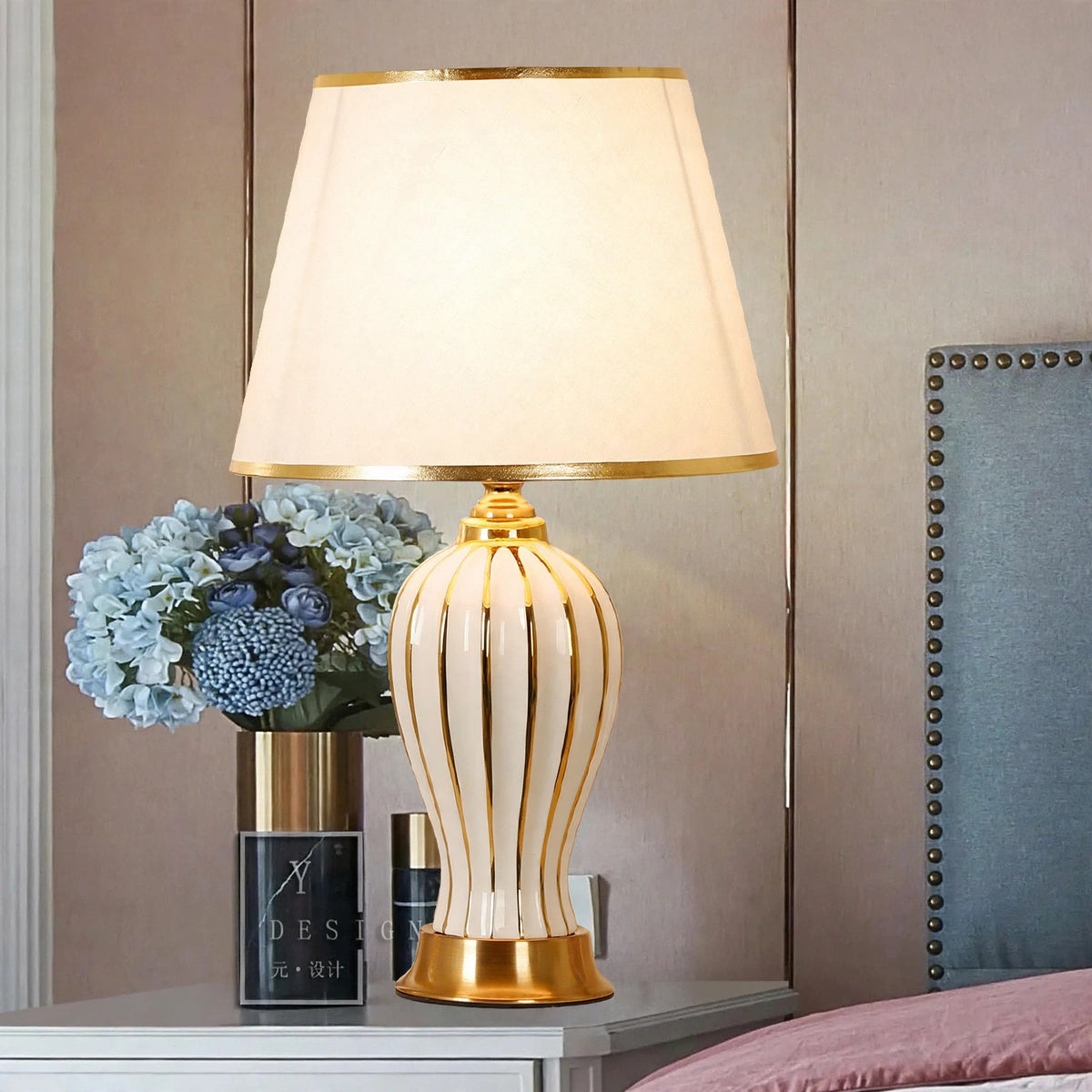 White ceramic table lamp with gold trim on a bedside table with hydrangea flowers, lit