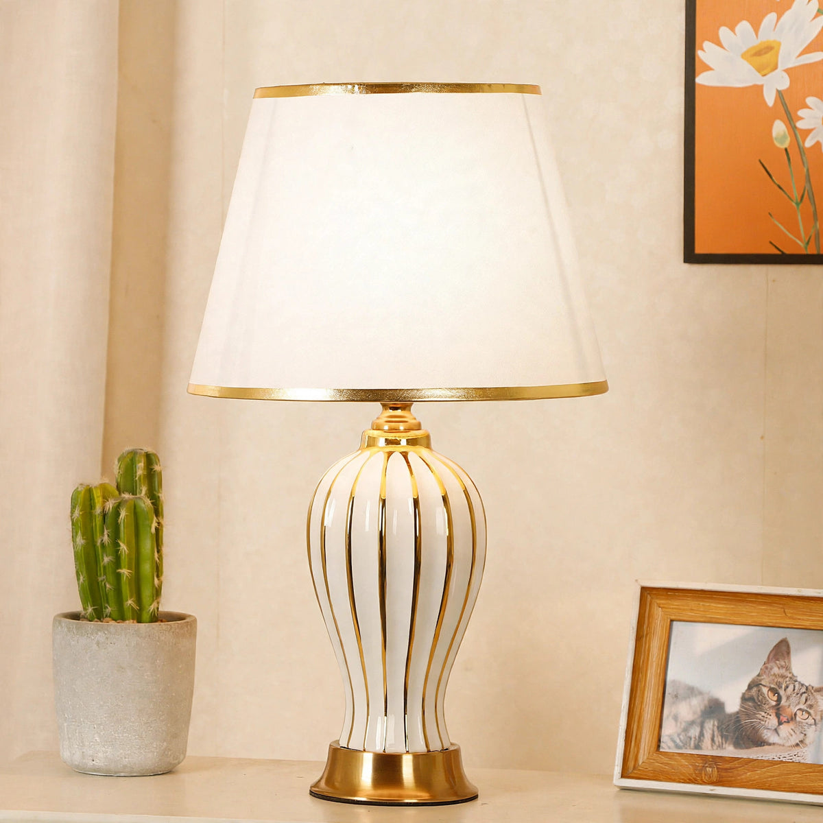 White ceramic table lamp lit on a side table with a potted cactus and framed photo