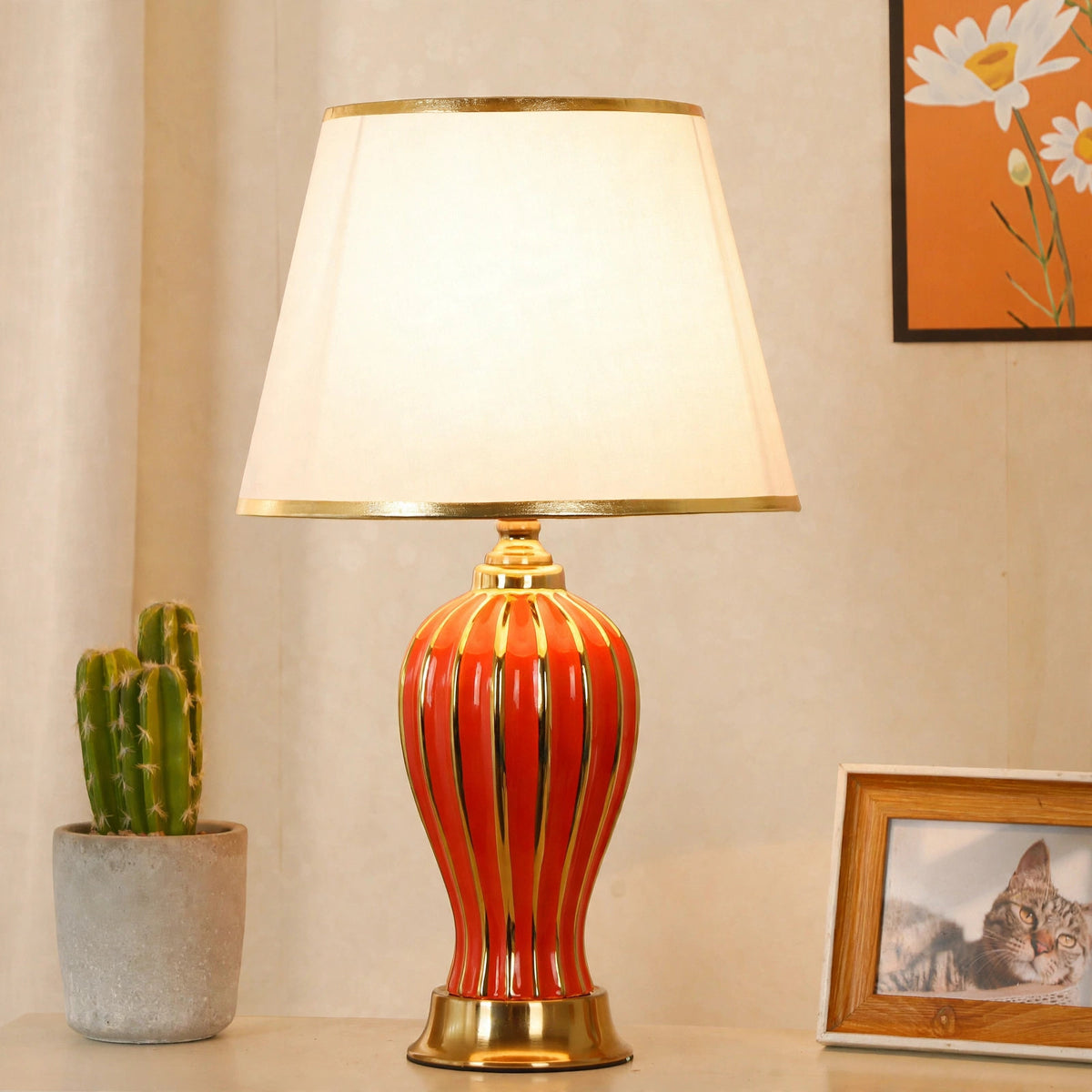 Red ceramic table lamp lit on a side table with a potted cactus and framed photo